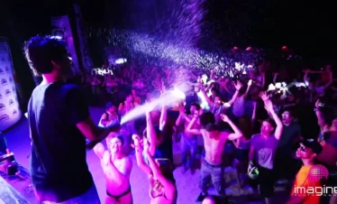 A crowd at a lively outdoor concert in Punta Cana dances and cheers as a performer sprays water over the audience. Colorful lights illuminate the scene, and many shirtless attendees revel in the energetic festival atmosphere.