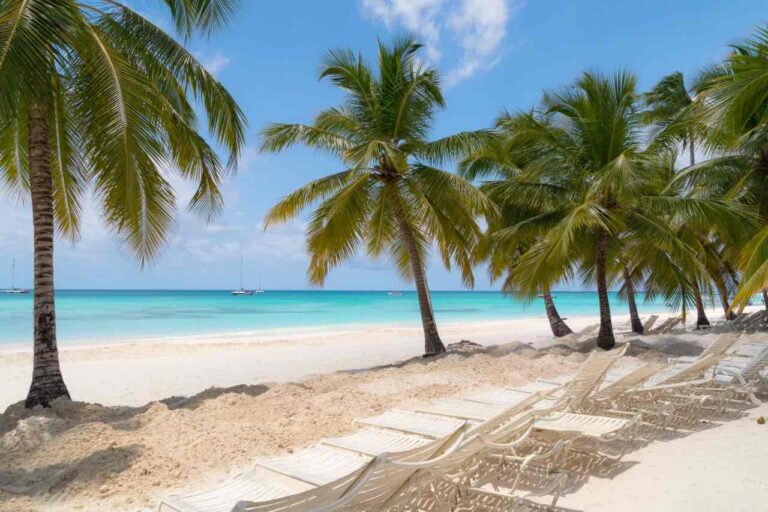 Rows of empty white lounge chairs sit on a sandy beach shaded by palm trees. The turquoise ocean is calm under a blue sky with scattered clouds. A few sailboats float on the water in the distance, creating a tranquil, tropical scene.