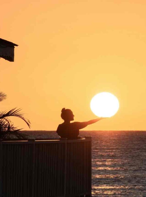 A person stands on a balcony by the sea at sunset, appearing to hold the sun in their outstretched hand. Palm leaves and part of a building are visible on the left, with the sky and water bathed in warm orange light.