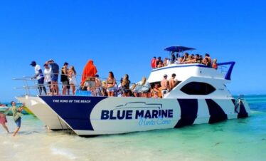 A crowded white and blue catamaran named “Blue Marine Punta Cana” is anchored in shallow, clear turquoise water. Dozens of people in swimsuits are on the boat and on the beach, enjoying sunny, blue-sky weather. Some walk along the sand; others relax onboard.