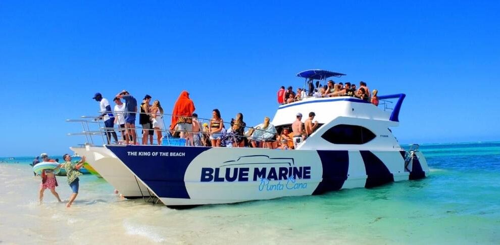 A crowded white and blue catamaran named “Blue Marine Punta Cana” is anchored in shallow, clear turquoise water. Dozens of people in swimsuits are on the boat and on the beach, enjoying sunny, blue-sky weather. Some walk along the sand; others relax onboard.