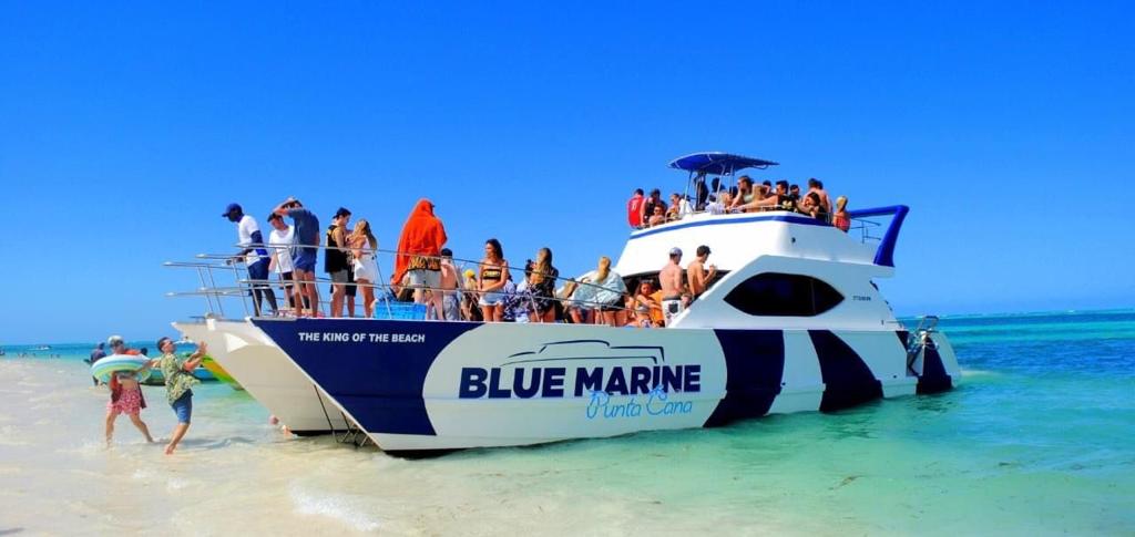 A crowded white and blue catamaran named “Blue Marine Punta Cana” is anchored in shallow, clear turquoise water. Dozens of people in swimsuits are on the boat and on the beach, enjoying sunny, blue-sky weather. Some walk along the sand; others relax onboard.