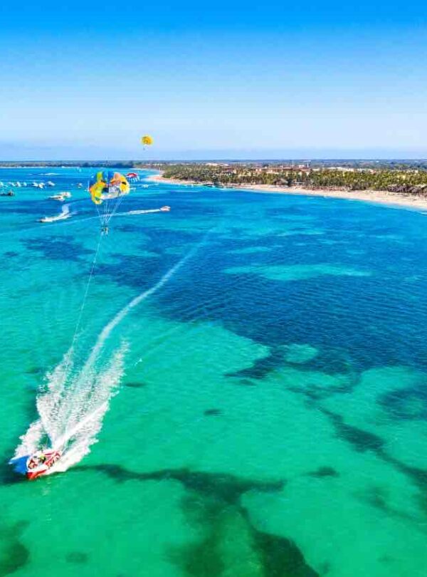 Aerial view of a speedboat pulling people on a yellow parasail over clear turquoise ocean water near a sandy beach with palm trees. Other boats are visible in the distance under a bright blue sky.