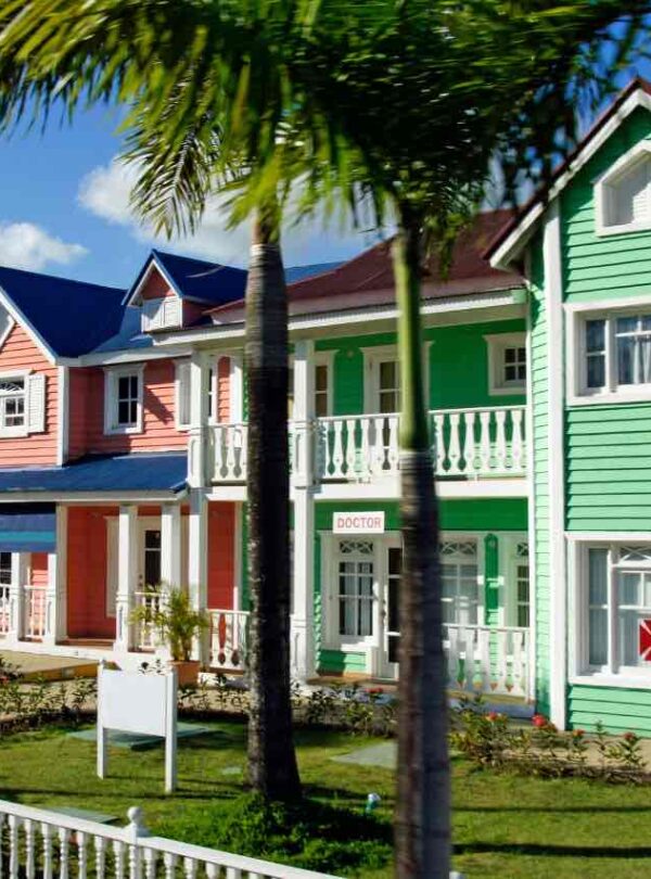 A row of colorful, two-story wooden houses with porches and white railings stands beside a white picket fence. Palm trees and flowers line the sidewalk. The houses are painted green, pink, and blue under a bright, sunny sky with scattered clouds.