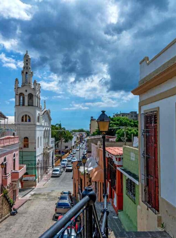 Colorful colonial buildings line a narrow street leading toward a white church with a tower. Cars are parked along the road. A wrought-iron balcony and classic street lamp feature in the foreground. Blue sky with dramatic clouds overhead.
