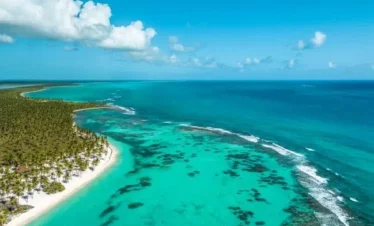Aerial view of Saona Island’s tropical beach with turquoise water, white sand, and palm trees lining the shore. The clear sea reveals coral reefs, while the vip sky above shines bright blue with scattered clouds.