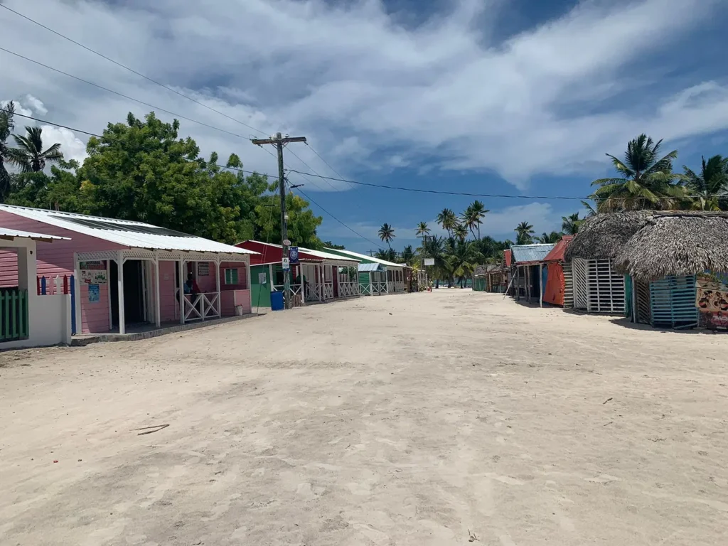 A wide sandy street on Saona Island, lined with colorful beach huts and shops, sits empty under a bright blue sky. Palm trees and lush greenery border the scene, creating a tropical, laid-back atmosphere with VIP vibes.