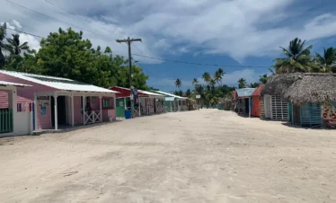 A wide sandy street on Saona Island, lined with colorful beach huts and shops, sits empty under a bright blue sky. Palm trees and lush greenery border the scene, creating a tropical, laid-back atmosphere with VIP vibes.