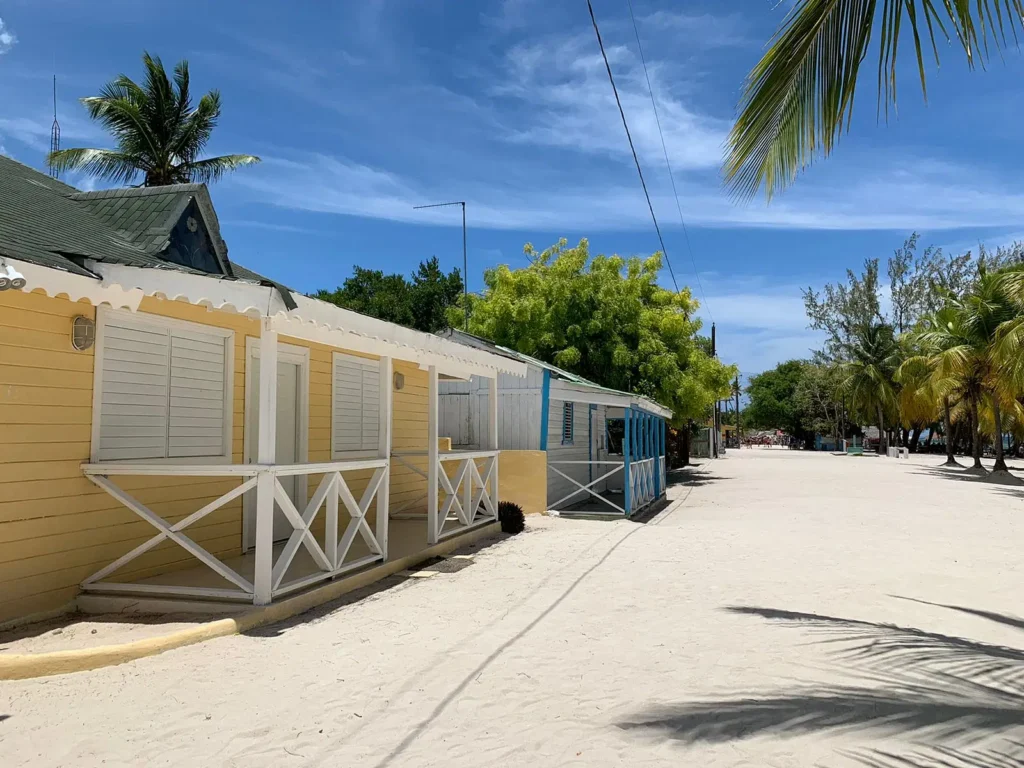 A sandy street lined with colorful wooden beach cabins, mostly painted yellow and blue, with white railings and shutters—captures the laid-back charm of a Saona Island beach, where palm trees and a clear sky complete the tropical vibe.