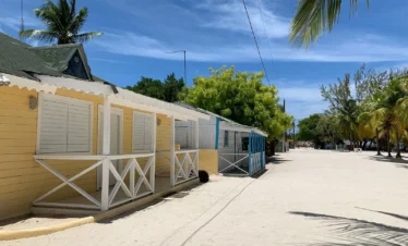 A sandy street lined with colorful wooden beach cabins, mostly painted yellow and blue, with white railings and shutters—captures the laid-back charm of a Saona Island beach, where palm trees and a clear sky complete the tropical vibe.