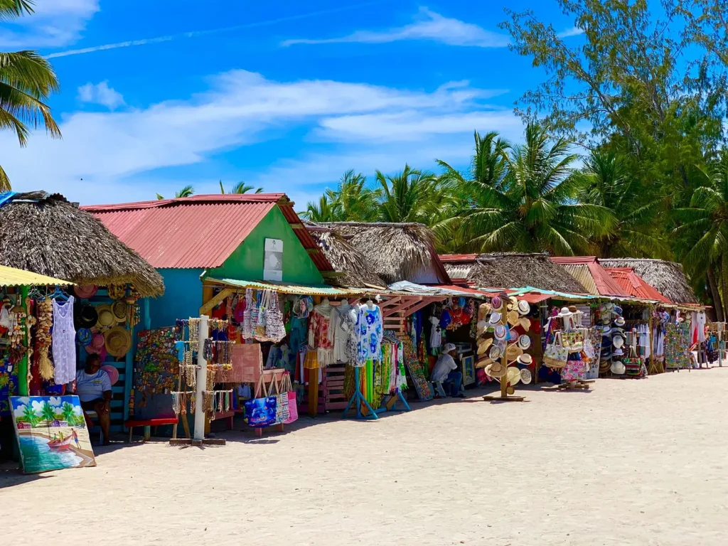 Colorful market stalls with red and green roofs line a sandy street on Saona Island, displaying hats, clothes, paintings, and crafts. Palm trees and blue sky in the background create a tropical atmosphere as people browse or relax at this beachside scene.