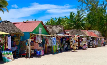 Colorful market stalls with red and green roofs line a sandy street on Saona Island, displaying hats, clothes, paintings, and crafts. Palm trees and blue sky in the background create a tropical atmosphere as people browse or relax at this beachside scene.