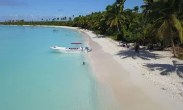 A tropical Saona Island beach with soft white sand, turquoise water, and palm trees. A small motorboat with a red canopy floats nearby as people relax and swim in the clear shallows—perfect for a Vip Tour of 3 Beach paradise under blue skies.