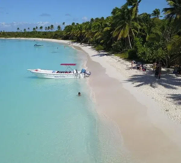 A tropical Saona Island beach with soft white sand, turquoise water, and palm trees. A small motorboat with a red canopy floats nearby as people relax and swim in the clear shallows—perfect for a Vip Tour of 3 Beach paradise under blue skies.