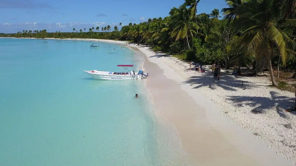 A tropical Saona Island beach with soft white sand, turquoise water, and palm trees. A small motorboat with a red canopy floats nearby as people relax and swim in the clear shallows—perfect for a Vip Tour of 3 Beach paradise under blue skies.
