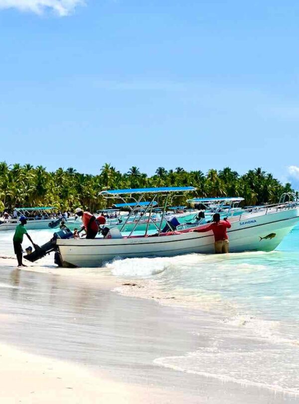 Two motorboats with people onboard rest in shallow turquoise water near a tropical sandy beach lined with palm trees. A man stands next to one boat, touching its motor, while the sky is mostly clear with a few clouds.