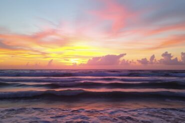 A vivid sunset over the ocean, with colorful clouds in shades of pink, orange, and yellow reflecting on gentle waves. The sea appears calm, and the horizon is visible, creating a peaceful and serene atmosphere.