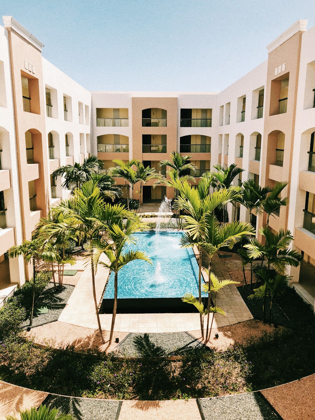 En el patio de un moderno edificio de Punta Cana (República Dominicana), con paredes de color crema y beige, hay una piscina rectangular con una fuente central, rodeada de palmeras y senderos ajardinados iluminados por el sol. Tres balcones dan a este sereno rincón.