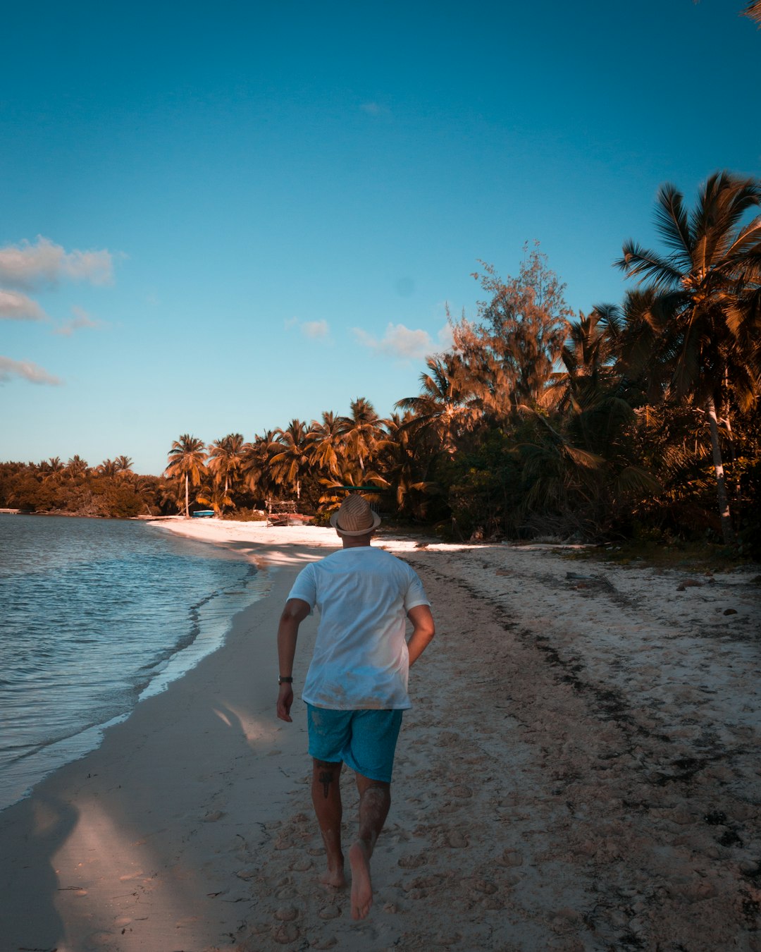 Un hombre con camiseta blanca y pantalones cortos azules camina descalzo por una playa de arena de Punta Cana bordeada de palmeras. El sol proyecta un cálido resplandor, el océano en calma queda a la izquierda y el cielo azul despejado crea un escenario perfecto para los paquetes de vacaciones en la República Dominicana.
