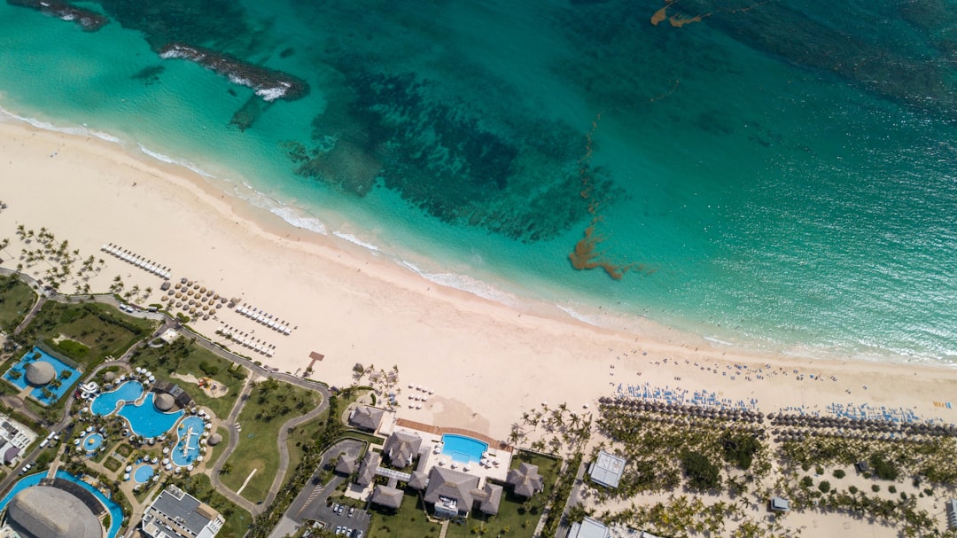 Vista aérea de una playa tropical de Punta Cana con agua turquesa, arena blanca y suaves olas. En tierra hay edificios de complejos turísticos, piscinas, palmeras e hileras de sombrillas, creando un ambiente relajante de vacaciones en la República Dominicana.